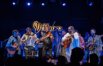 Seis músicos en el escenario: un guitarrista con poncho, un acordeonista, un violinista, una cantante y otros con guitarra y saxofón.
