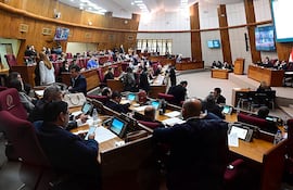 Diputados en salón legislativo, algunos escribiendo y conversando, rodeados de un ambiente formal y pantallas en el fondo.
