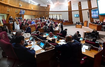 Diputados en salón legislativo, algunos escribiendo y conversando, rodeados de un ambiente formal y pantallas en el fondo.