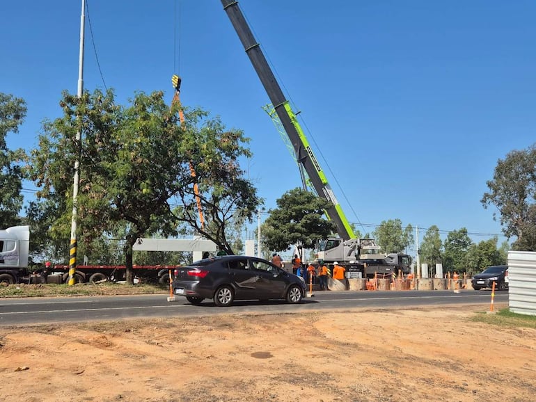 Camión y vehículo gris en obra vial, con trabajadores en camisetas naranjas operando grúa bajo cielo despejado.