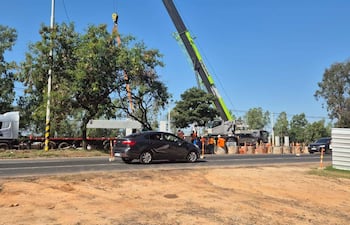 Camión y vehículo gris en obra vial, con trabajadores en camisetas naranjas operando grúa bajo cielo despejado.