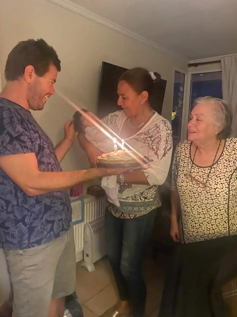 Mujer sonriente sostiene un pastel de cumpleaños mientras un hombre acaricia la cabeza de un bebé en brazos. Ambiente festivo.