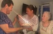 Mujer sonriente sostiene un pastel de cumpleaños mientras un hombre acaricia la cabeza de un bebé en brazos. Ambiente festivo.