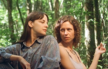 Dos mujeres sentadas en la jungla, una con cabello lacio y oscuro y otra con cabello rizado, sosteniendo un cigarrillo.