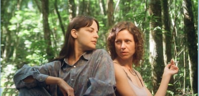 Dos mujeres sentadas en la jungla, una con cabello lacio y oscuro y otra con cabello rizado, sosteniendo un cigarrillo.