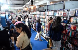 Hombre con abrigo gris habla con mujer, mientras tres mujeres revisan libros en un ambiente iluminado y colorido.