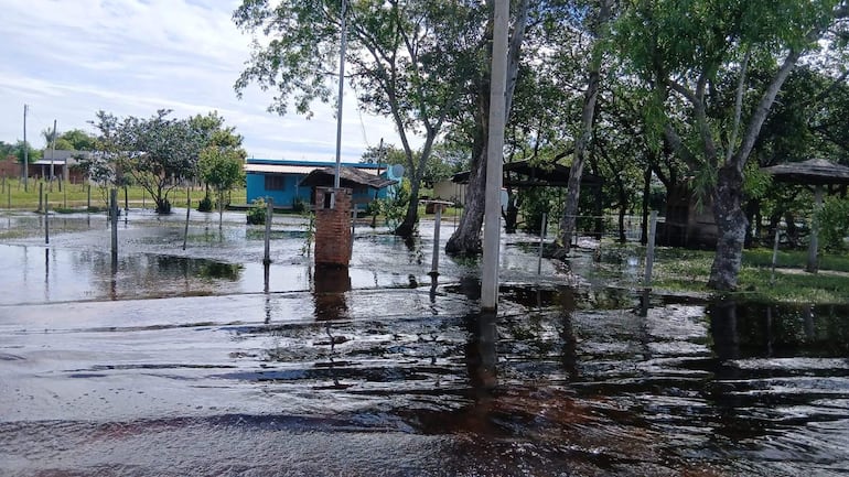Área inundada con agua cubriendo el suelo, rodeada de árboles y edificios, sin personas presentes.