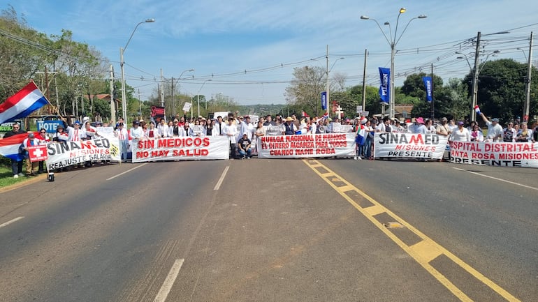 Grupo de médicos en camisetas blancas y batas, sosteniendo pancartas en la calle, expresando su protesta con seriedad.