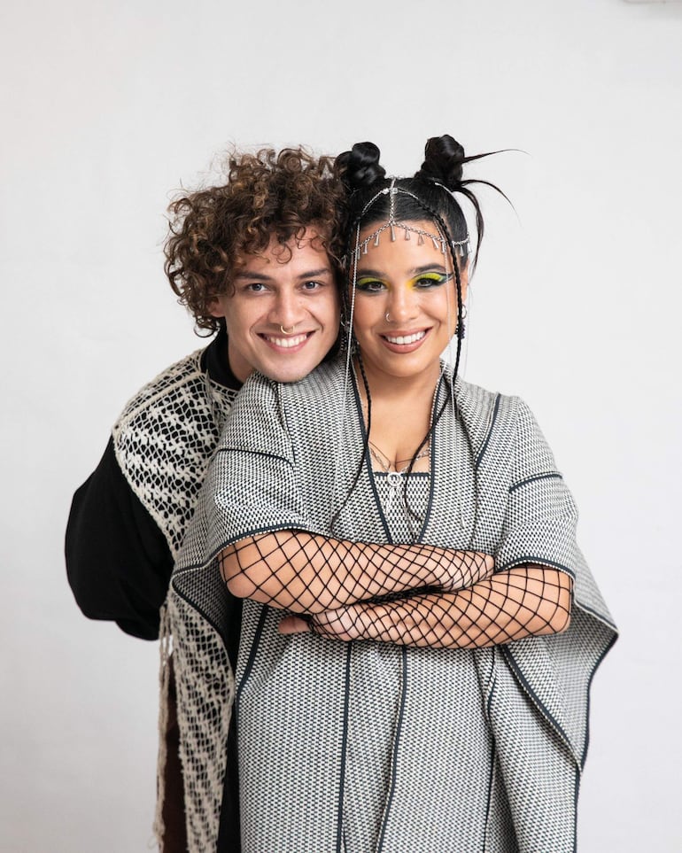 Dos jóvenes sonrientes, una con moños altos y atuendo blanco y negro, la otra con cabello rizado y abrigo claro, en un fondo neutro.