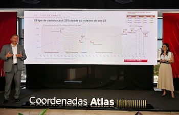 Patricia Goto en vestido blanco junto a un hombre en traje gris que presenta gráficos sobre economía en un ambiente moderno.