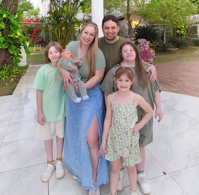Mujer sosteniendo un bebé, sonriente, rodeada de un hombre y tres niños en un entorno natural.