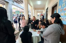 Mujer rubia con micrófono habla mientras tres personas observan y escriben en una mesa con documentos en un ambiente iluminado.