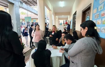 Mujer rubia con micrófono habla mientras tres personas observan y escriben en una mesa con documentos en un ambiente iluminado.