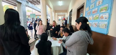 Mujer rubia con micrófono habla mientras tres personas observan y escriben en una mesa con documentos en un ambiente iluminado.