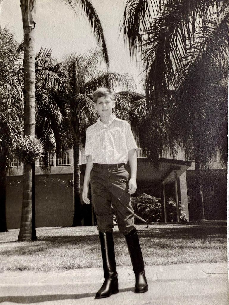 Niño con camisa blanca y pantalones cortos, sonriente entre palmeras, en un ambiente soleado y tranquilo.