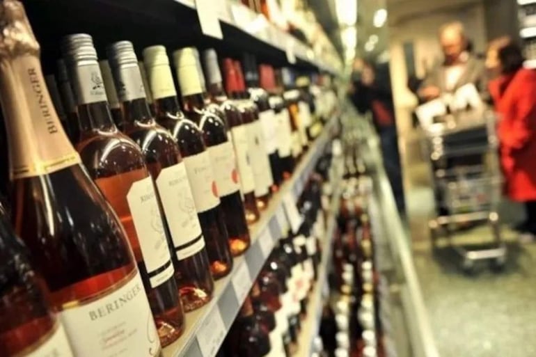 Hombre canoso en abrigo gris observa vinos, mientras mujer en abrigo rojo conversa con otro cliente en la tienda.