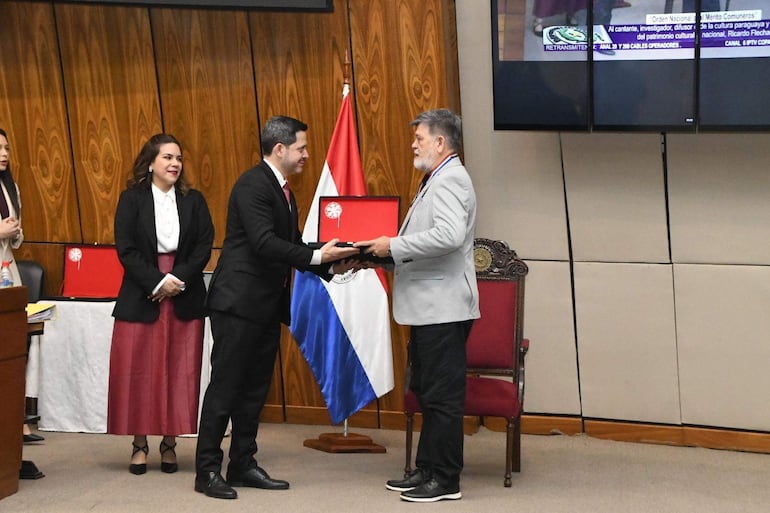 Ricardo Flecha Hermosa en traje claro con lazo, mientras un hombre de traje oscuro le entrega un premio en un entorno ceremonial.