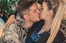 Pareja en un entorno íntimo, hombre con chaqueta destacada y mujer con blazer negro y camisa azul, ambos sonriendo mientras se besan.