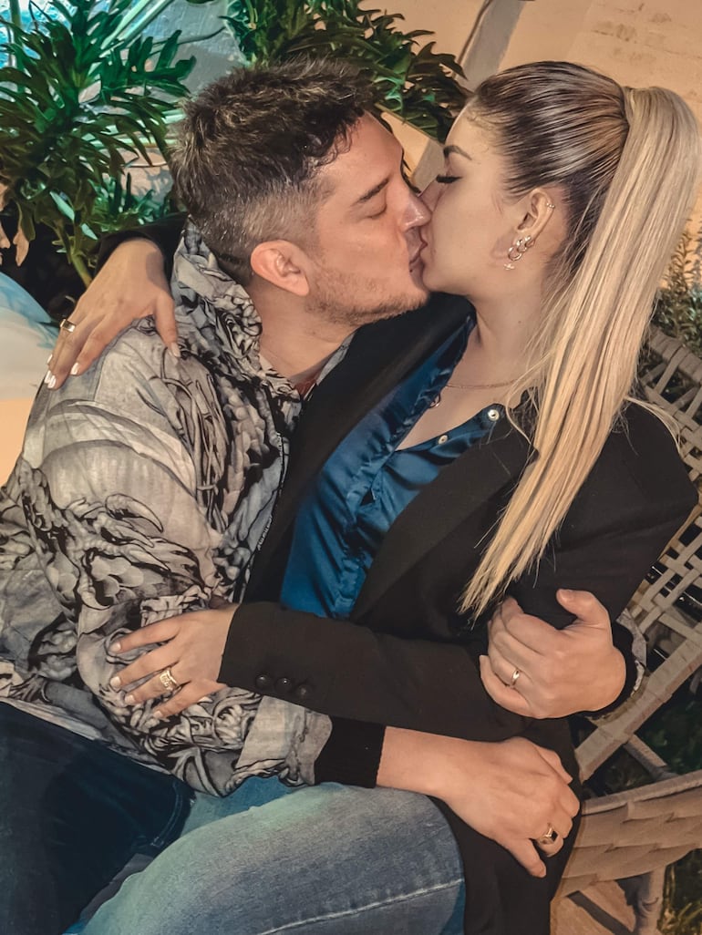 Pareja en un entorno íntimo, hombre con chaqueta destacada y mujer con blazer negro y camisa azul, ambos sonriendo mientras se besan.