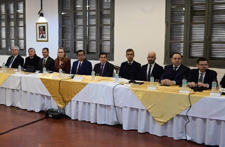 Diez personas en trajes oscuros discuten en una mesa rectangular. Ambiente formal con ventanales y fotografía en la pared.