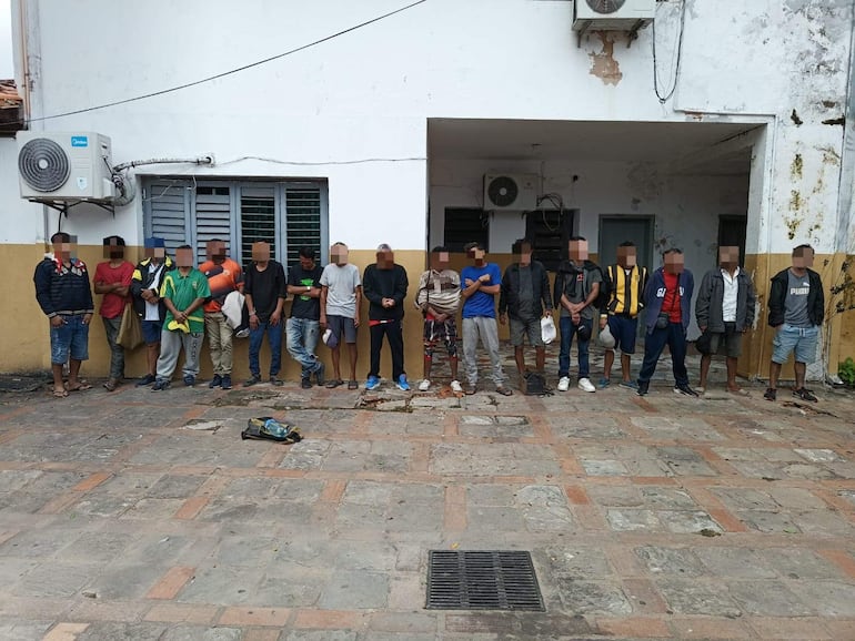 Grupo de 16 personas alineadas contra una pared, vestidas variadamente, esperando en un ambiente exterior con baldosas y paredes de yeso.