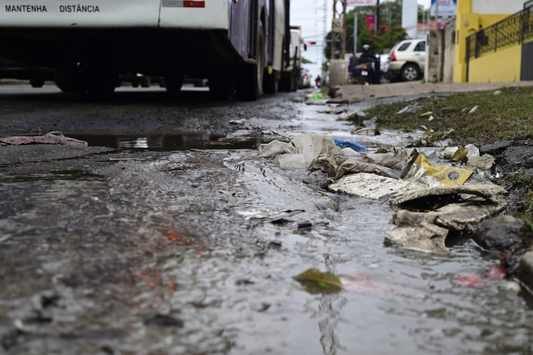 Calle con zanja llena de basura y agua estancada, sin personas presentes, vehículos al fondo.
