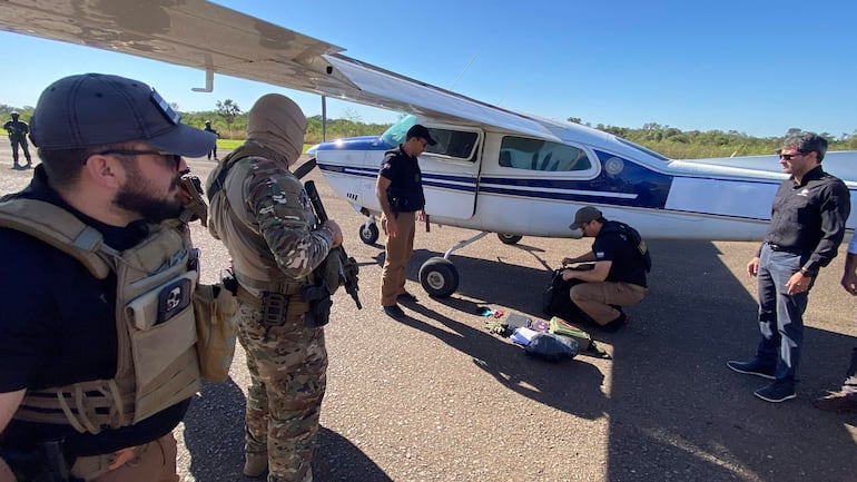 Cuatro hombres en ropa táctica observan una aeronave blanca y azul en el aeródromo con un cielo despejado al fondo.