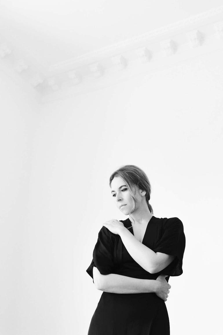 Rocío Márquez con vestido negro en un fondo minimalista, abrazándose los brazos en postura contemplativa.