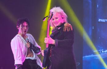 Lenny Jay con camiseta blanca, cantando fervientemente, y Jennifer Batten sonriente con guitarra y abrigo negro, sobre un escenario iluminado.