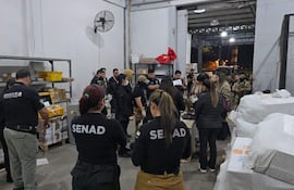 Mujer del Senad habla con documentos en mano, rodeada de otros, en un ambiente de almacenamiento iluminado artificialmente.