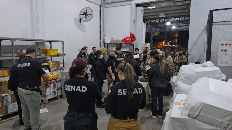 Mujer del Senad habla con documentos en mano, rodeada de otros, en un ambiente de almacenamiento iluminado artificialmente.
