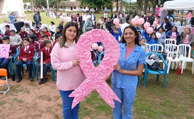 Ángela Cáceres sonríe con suéter rosa, mientras Análisis Espínola sostiene un lazo rosa, rodeadas de niños en un parque.