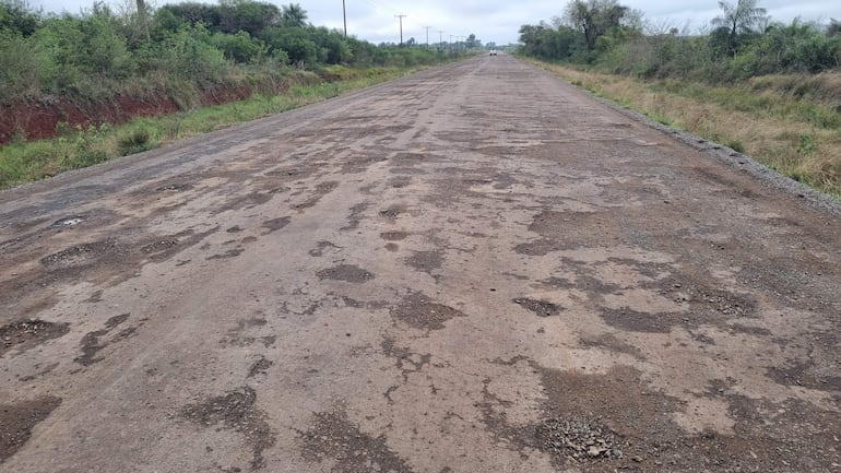 Carretera deteriorada con baches, rodeada de vegetación y cielo nublado, sin tráfico visible.
