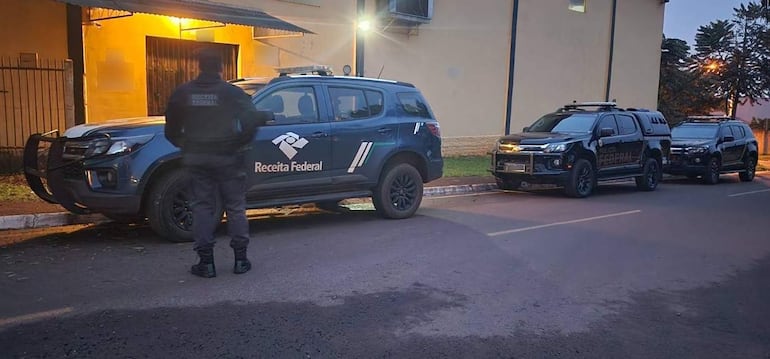 Agente de uniforme oscuro de Policía Federal conversa en un entorno urbano con vehículos rotulados de Receita Federal.