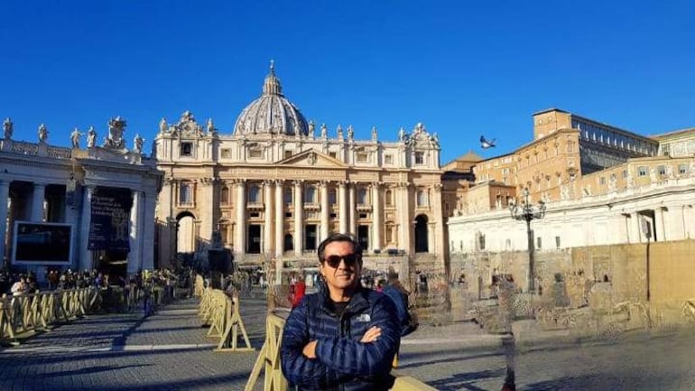 Turista con gafas de sol y chaqueta oscura, sonriente y con los brazos cruzados, en la Plaza de San Pedro con la Basílica de fondo.