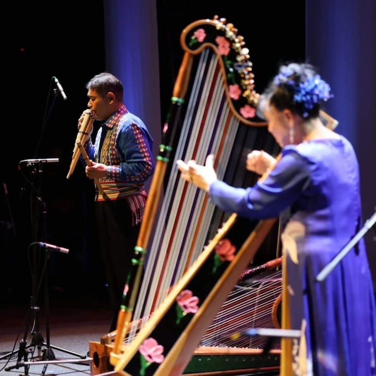 Músico hombre en atuendo colorido tocando flauta y mujer en traje azul tocando arpa en un escenario iluminado.