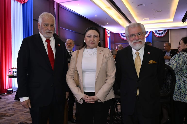 Hombre mayor con barba gris y corbata roja, mujer en blazer claro y hombre con barba blanca en evento decorado con colores patrios.