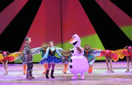 Mujer en vestido vibrante interactúa con un muñeco de nieve, mientras joven observa en el escenario lleno de colores festivos.