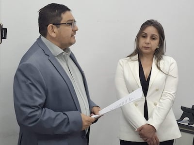 Hombre en traje gris holding un documento, mientras una mujer con abrigo blanco escucha atentamente.