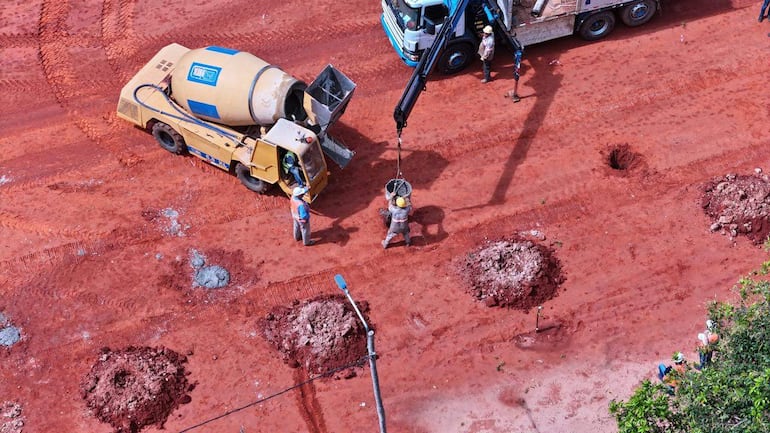 Cinco trabajadores con cascos, alrededor de hormigonera amarilla, operan grúa en construcción con tierra roja y agujeros.