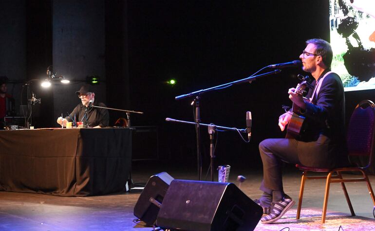 Kevin Johansen, con traje oscuro y gafas, toca la guitarra mientras Liniers, en camiseta negra y gorra, lo asiste desde una mesa.