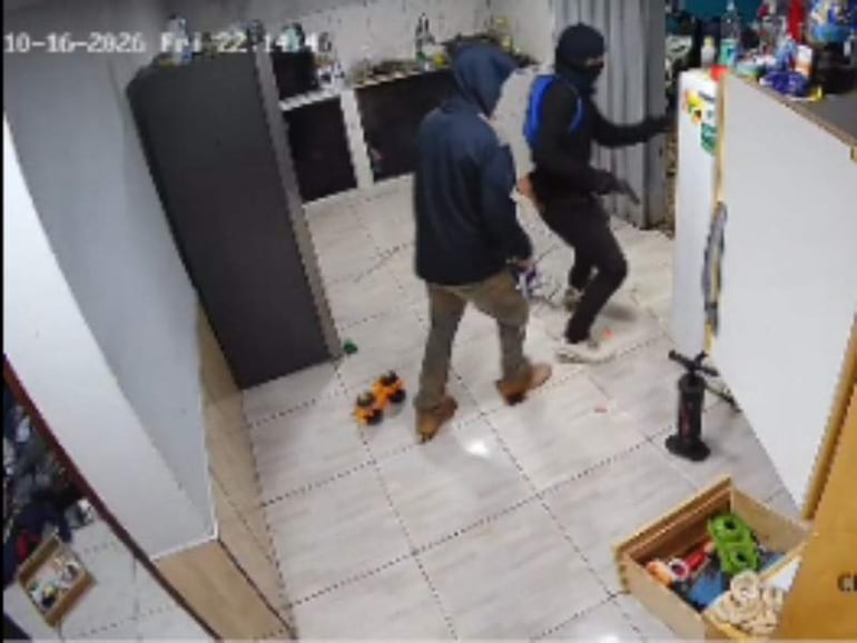 Dos hombres en vivienda con rostros cubiertos, uno de pie con chaqueta oscura y el otro en atuendo negro con máscara.