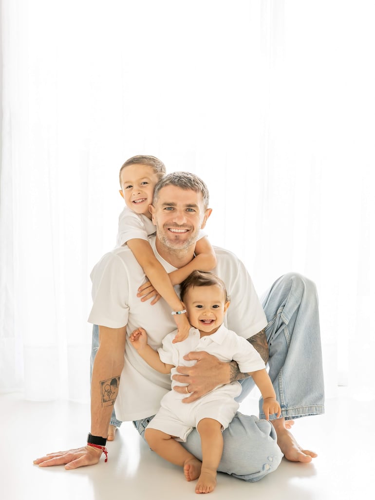 Pablo Vegetti en camiseta blanca sonríe mientras sostiene a un niño pequeño y otro niño está sobre su espalda, todo en un fondo blanco.