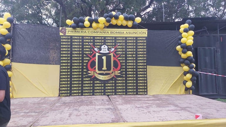 Escenario decorado con panel que dice 'PRIMERA COMPAÑÍA BOMBA ASUNCIÓN', globos negros y amarillos, ambiente al aire libre.