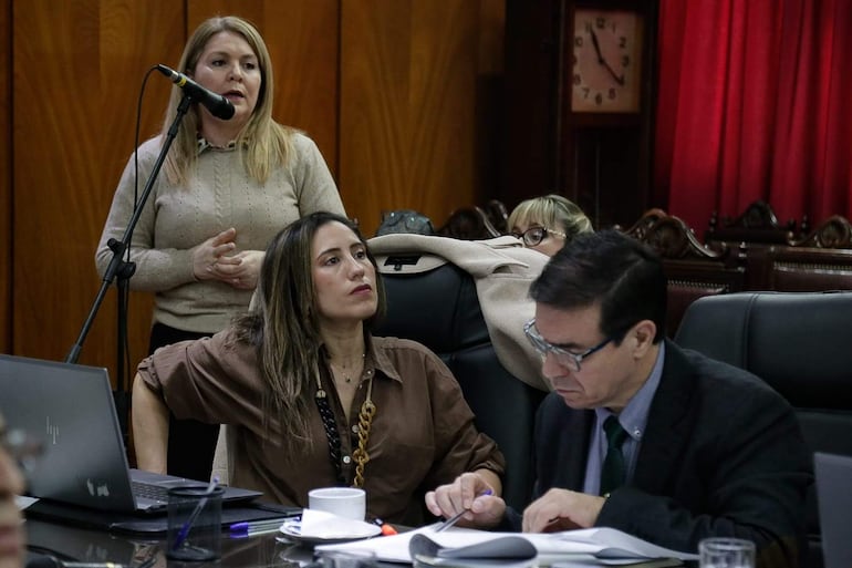 Bettina Albertini habla al micrófono con vestimenta clara, mientras otra mujer observa y un hombre toma notas.