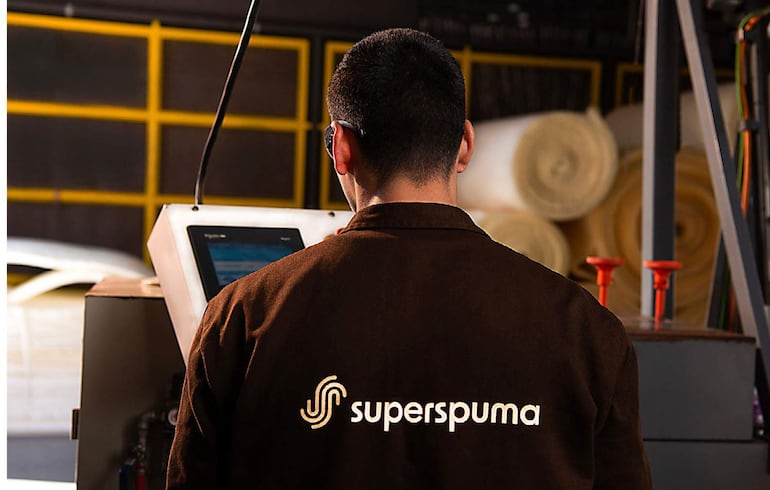 Hombre de espaldas con chaqueta marrón y logotipo de superspuma, operando máquina en taller con rollos de material.