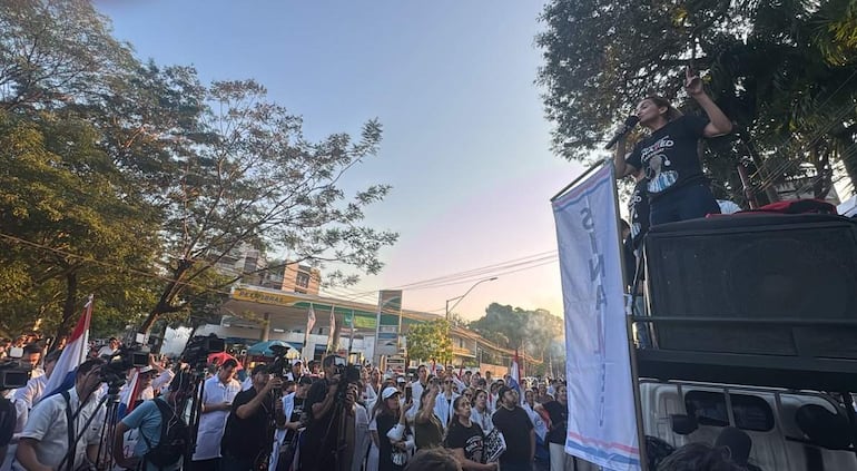 Rossana González en camiseta negra sostiene un micrófono desde un vehículo, rodeada de una multitud variada, algunos aplaudiendo.