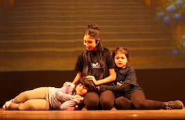 Niña de cabello largo en camiseta negra interactúa con dos compañeras en un escenario teatral. Ambiente cálido y de entretenimiento.