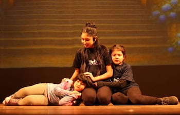 Niña de cabello largo en camiseta negra interactúa con dos compañeras en un escenario teatral. Ambiente cálido y de entretenimiento.
