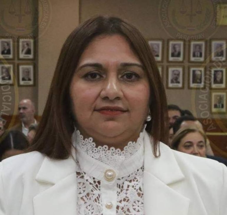 Mujer de mediana edad con cabello largo y lacio, en blusa clara y abrigo blanco, observa a la cámara en un entorno formal.